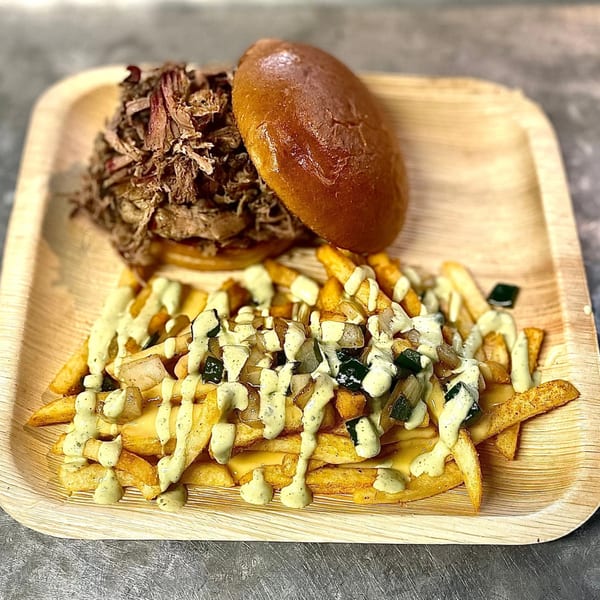 Pulled pork sandwich on a brioche bun with loaded fries topped with sauce, onions, and zucchini.