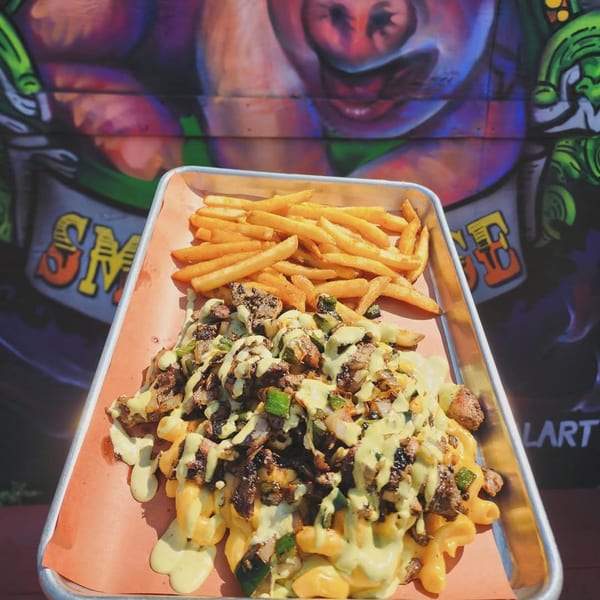 BBQ mac and cheese with meat, peppers, onions, and fries on a tray, in front of a pig mural.