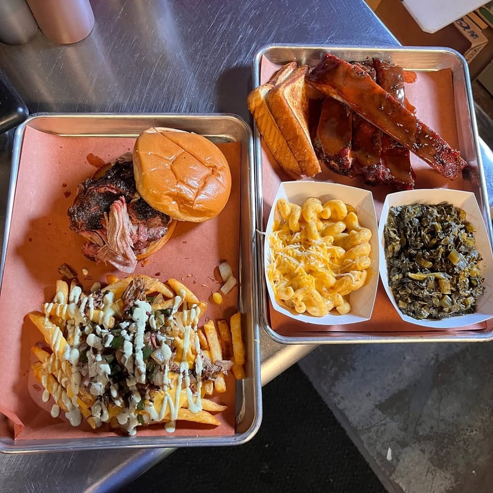 Two trays of Butt Hutt Smokehouse BBQ: pulled pork sandwich, loaded fries, ribs, mac and cheese, and collard greens.